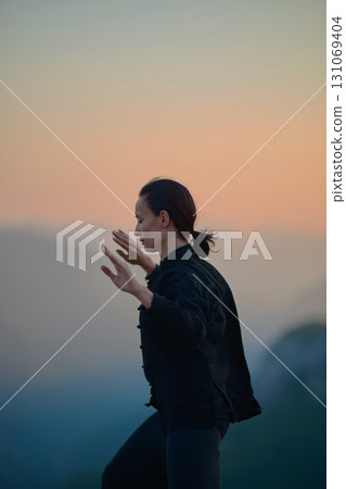 Woman practicing Tai Chi in traditional black outfit on mountain at sunrise, arms outstretched in meditation pose focusing on balance, energy, and mindfulness in nature. 131069404