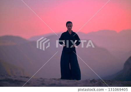 Woman practicing Tai Chi in traditional black outfit on mountain at sunrise, arms outstretched in meditation pose focusing on balance, energy, and mindfulness in nature. 131069405