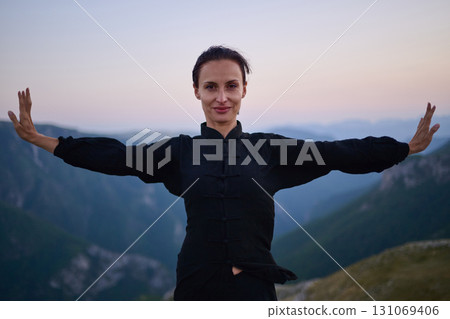 Woman practicing Tai Chi in traditional black outfit on mountain at sunrise, arms outstretched in meditation pose focusing on balance, energy, and mindfulness in nature. Woman practicing Tai Chi in traditional black outfit on mountain at sunrise, arms outstretched in meditation pose focusing on balance, energy, and mindfulness in nature. 131069406