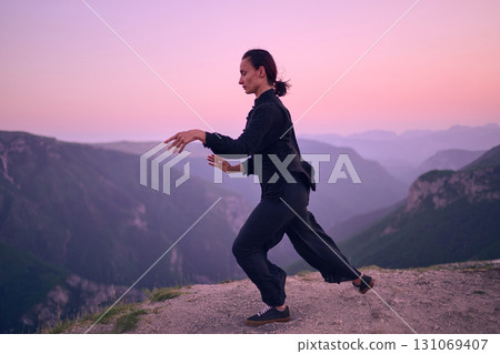 Woman practicing Tai Chi in traditional black outfit on mountain at sunrise, arms outstretched in meditation pose focusing on balance, energy, and mindfulness in nature. 131069407