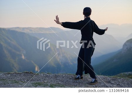 Woman practicing Tai Chi in traditional black outfit on mountain at sunrise, arms outstretched in meditation pose focusing on balance, energy, and mindfulness in nature. 131069416