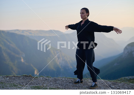 Woman practicing Tai Chi in traditional black outfit on mountain at sunrise, arms outstretched in meditation pose focusing on balance, energy, and mindfulness in nature. 131069417