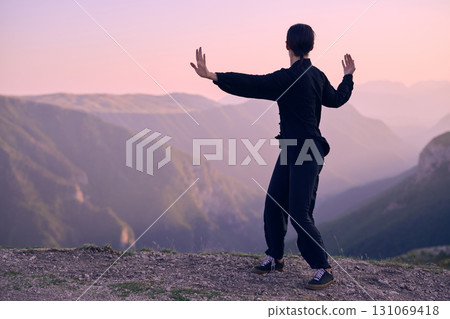 Woman practicing Tai Chi in traditional black outfit on mountain at sunrise, arms outstretched in meditation pose focusing on balance, energy, and mindfulness in nature. 131069418