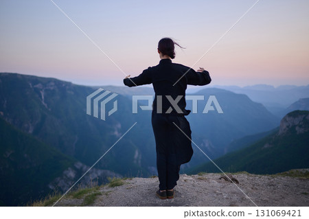 Woman practicing Tai Chi in traditional black outfit on mountain at sunrise, arms outstretched in meditation pose focusing on balance, energy, and mindfulness in nature. 131069421