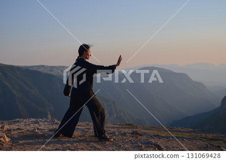 Woman practicing Tai Chi in traditional black outfit on mountain at sunrise, arms outstretched in meditation pose focusing on balance, energy, and mindfulness in nature. Woman practicing Tai Chi in traditional black outfit on mountain at sunrise, arms outstretched in meditation pose focusing on balance, energy, and mindfulness in nature. 131069428
