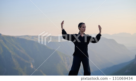 Woman practicing Tai Chi in traditional black outfit on mountain at sunrise, arms outstretched in meditation pose focusing on balance, energy, and mindfulness in nature. Woman practicing Tai Chi in traditional black outfit on mountain at sunrise, arms outstretched in meditation pose focusing on balance, energy, and mindfulness in nature. 131069434