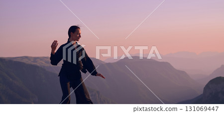 Woman practicing Tai Chi in traditional black outfit on mountain at sunrise, arms outstretched in meditation pose focusing on balance, energy, and mindfulness in nature. 131069447
