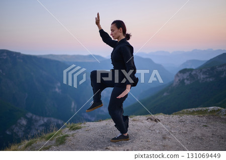 Woman practicing Tai Chi in traditional black outfit on mountain at sunrise, arms outstretched in meditation pose focusing on balance, energy, and mindfulness in nature. 131069449