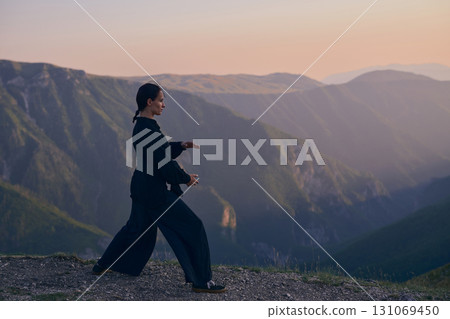 Woman practicing Tai Chi in traditional black outfit on mountain at sunrise, arms outstretched in meditation pose focusing on balance, energy, and mindfulness in nature. 131069450