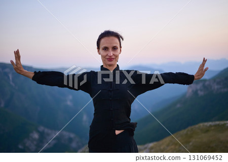 Woman practicing Tai Chi in traditional black outfit on mountain at sunrise, arms outstretched in meditation pose focusing on balance, energy, and mindfulness in nature. 131069452