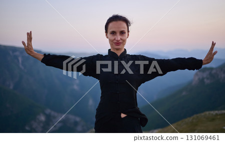 Woman practicing Tai Chi in traditional black outfit on mountain at sunrise, arms outstretched in meditation pose focusing on balance, energy, and mindfulness in nature. 131069461