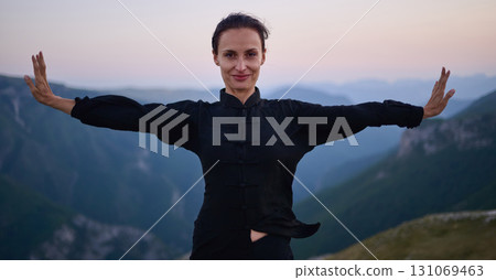 Woman practicing Tai Chi in traditional black outfit on mountain at sunrise, arms outstretched in meditation pose focusing on balance, energy, and mindfulness in nature. 131069463