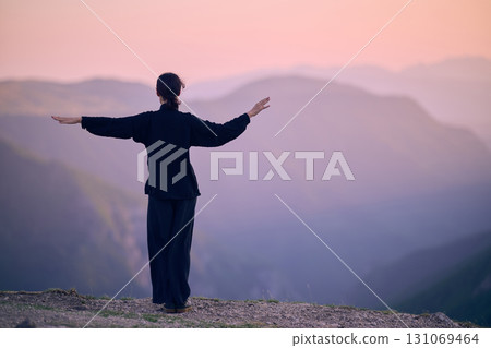 Woman practicing Tai Chi in traditional black outfit on mountain at sunrise, arms outstretched in meditation pose focusing on balance, energy, and mindfulness in nature. 131069464