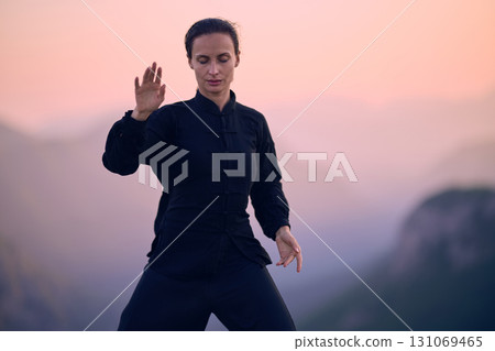 Woman practicing Tai Chi in traditional black outfit on mountain at sunrise, arms outstretched in meditation pose focusing on balance, energy, and mindfulness in nature. 131069465