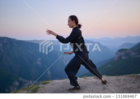 Woman practicing Tai Chi in traditional black outfit on mountain at sunrise, arms outstretched in meditation pose focusing on balance, energy, and mindfulness in nature. 131069467