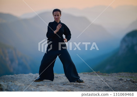 Woman practicing Tai Chi in traditional black outfit on mountain at sunrise, arms outstretched in meditation pose focusing on balance, energy, and mindfulness in nature. 131069468