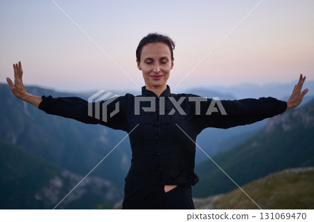 Woman practicing Tai Chi in traditional black outfit on mountain at sunrise, arms outstretched in meditation pose focusing on balance, energy, and mindfulness in nature. Woman practicing Tai Chi in traditional black outfit on mountain at sunrise, arms outstretched in meditation pose focusing on balance, energy, and mindfulness in nature. 131069470