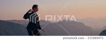 Woman practicing Tai Chi in traditional black outfit on mountain at sunrise, arms outstretched in meditation pose focusing on balance, energy, and mindfulness in nature. 131069471
