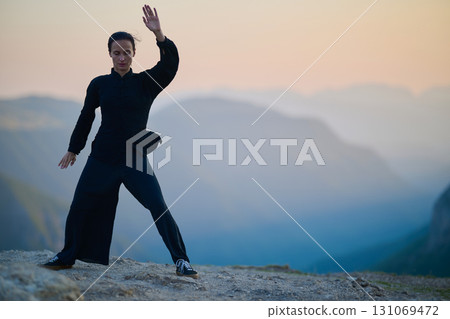 Woman practicing Tai Chi in traditional black outfit on mountain at sunrise, arms outstretched in meditation pose focusing on balance, energy, and mindfulness in nature. 131069472