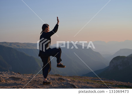 Woman practicing Tai Chi in traditional black outfit on mountain at sunrise, arms outstretched in meditation pose focusing on balance, energy, and mindfulness in nature. 131069473