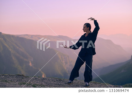 Woman practicing Tai Chi in traditional black outfit on mountain at sunrise, arms outstretched in meditation pose focusing on balance, energy, and mindfulness in nature. 131069474