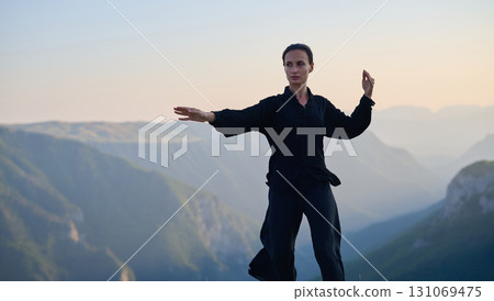 Woman practicing Tai Chi in traditional black outfit on mountain at sunrise, arms outstretched in meditation pose focusing on balance, energy, and mindfulness in nature. 131069475
