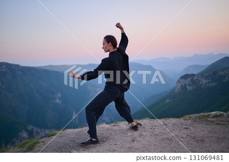 Woman practicing Tai Chi in traditional black outfit on mountain at sunrise, arms outstretched in meditation pose focusing on balance, energy, and mindfulness in nature. Woman practicing Tai Chi in traditional black outfit on mountain at sunrise, arms outstretched in meditation pose focusing on balance, energy, and mindfulness in nature. 131069481