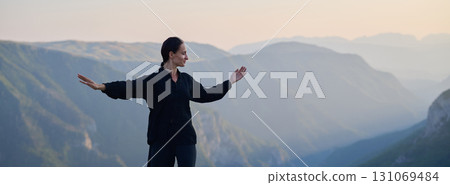 Woman practicing Tai Chi in traditional black outfit on mountain at sunrise, arms outstretched in meditation pose focusing on balance, energy, and mindfulness in nature. Woman practicing Tai Chi in traditional black outfit on mountain at sunrise, arms outstretched in meditation pose focusing on balance, energy, and mindfulness in nature. 131069484