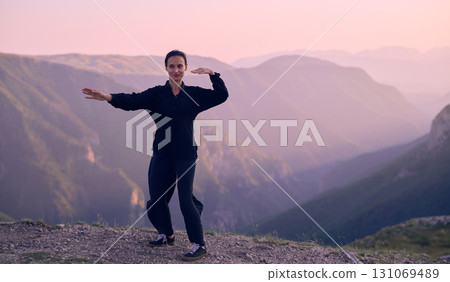 Woman practicing Tai Chi in traditional black outfit on mountain at sunrise, arms outstretched in meditation pose focusing on balance, energy, and mindfulness in nature. 131069489