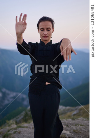 Woman practicing Tai Chi in traditional black outfit on mountain at sunrise, arms outstretched in meditation pose focusing on balance, energy, and mindfulness in nature. 131069491