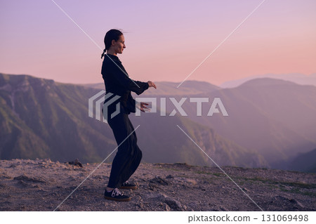 Woman practicing Tai Chi in traditional black outfit on mountain at sunrise, arms outstretched in meditation pose focusing on balance, energy, and mindfulness in nature. Woman practicing Tai Chi in traditional black outfit on mountain at sunrise, arms outstretched in meditation pose focusing on balance, energy, and mindfulness in nature. 131069498