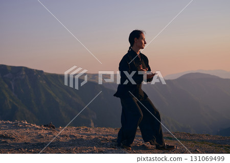 Woman practicing Tai Chi in traditional black outfit on mountain at sunrise, arms outstretched in meditation pose focusing on balance, energy, and mindfulness in nature. Woman practicing Tai Chi in traditional black outfit on mountain at sunrise, arms outstretched in meditation pose focusing on balance, energy, and mindfulness in nature. 131069499