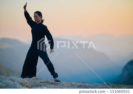 Woman practicing Tai Chi in traditional black outfit on mountain at sunrise, arms outstretched in meditation pose focusing on balance, energy, and mindfulness in nature. 131069501
