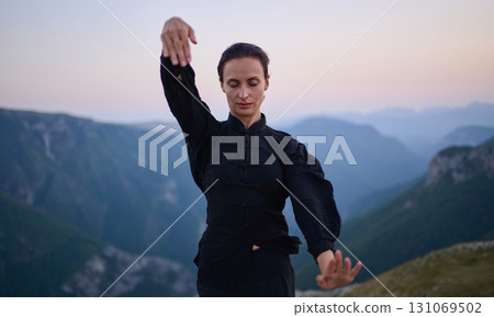 Woman practicing Tai Chi in traditional black outfit on mountain at sunrise, arms outstretched in meditation pose focusing on balance, energy, and mindfulness in nature. 131069502