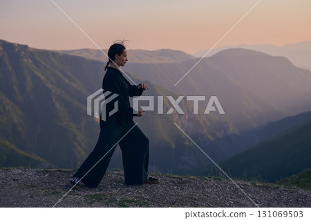 Woman practicing Tai Chi in traditional black outfit on mountain at sunrise, arms outstretched in meditation pose focusing on balance, energy, and mindfulness in nature. Woman practicing Tai Chi in traditional black outfit on mountain at sunrise, arms outstretched in meditation pose focusing on balance, energy, and mindfulness in nature. 131069503