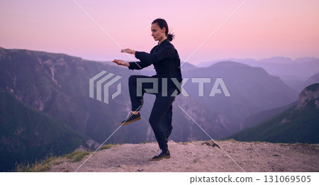 Woman practicing Tai Chi in traditional black outfit on mountain at sunrise, arms outstretched in meditation pose focusing on balance, energy, and mindfulness in nature. 131069505