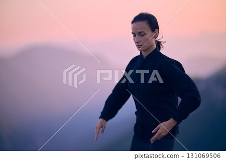 Woman practicing Tai Chi in traditional black outfit on mountain at sunrise, arms outstretched in meditation pose focusing on balance, energy, and mindfulness in nature. 131069506