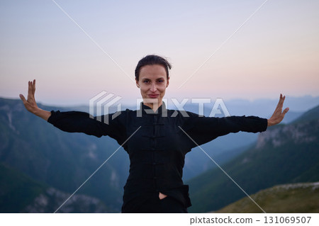 Woman practicing Tai Chi in traditional black outfit on mountain at sunrise, arms outstretched in meditation pose focusing on balance, energy, and mindfulness in nature. 131069507
