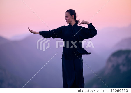 Woman practicing Tai Chi in traditional black outfit on mountain at sunrise, arms outstretched in meditation pose focusing on balance, energy, and mindfulness in nature. 131069510