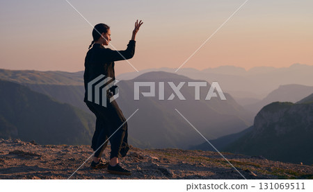 Woman practicing Tai Chi in traditional black outfit on mountain at sunrise, arms outstretched in meditation pose focusing on balance, energy, and mindfulness in nature. 131069511