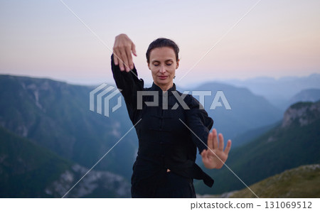 Woman practicing Tai Chi in traditional black outfit on mountain at sunrise, arms outstretched in meditation pose focusing on balance, energy, and mindfulness in nature. 131069512