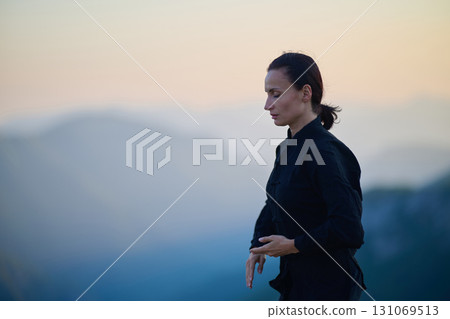 Woman practicing Tai Chi in traditional black outfit on mountain at sunrise, arms outstretched in meditation pose focusing on balance, energy, and mindfulness in nature. Woman practicing Tai Chi in traditional black outfit on mountain at sunrise, arms outstretched in meditation pose focusing on balance, energy, and mindfulness in nature. 131069513