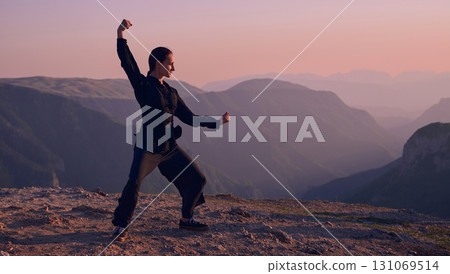 Woman practicing Tai Chi in traditional black outfit on mountain at sunrise, arms outstretched in meditation pose focusing on balance, energy, and mindfulness in nature. 131069514