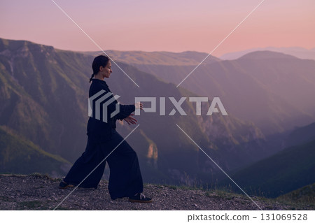 Woman practicing Tai Chi in traditional black outfit on mountain at sunrise, arms outstretched in meditation pose focusing on balance, energy, and mindfulness in nature. Woman practicing Tai Chi in traditional black outfit on mountain at sunrise, arms outstretched in meditation pose focusing on balance, energy, and mindfulness in nature. 131069528