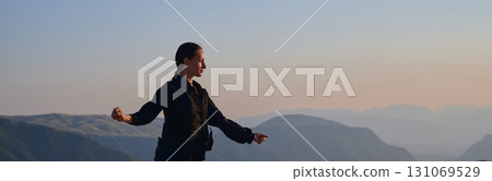 Woman practicing Tai Chi in traditional black outfit on mountain at sunrise, arms outstretched in meditation pose focusing on balance, energy, and mindfulness in nature. Woman practicing Tai Chi in traditional black outfit on mountain at sunrise, arms outstretched in meditation pose focusing on balance, energy, and mindfulness in nature. 131069529
