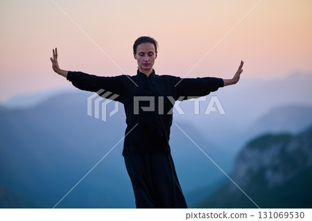 Woman practicing Tai Chi in traditional black outfit on mountain at sunrise, arms outstretched in meditation pose focusing on balance, energy, and mindfulness in nature. Woman practicing Tai Chi in traditional black outfit on mountain at sunrise, arms outstretched in meditation pose focusing on balance, energy, and mindfulness in nature. 131069530