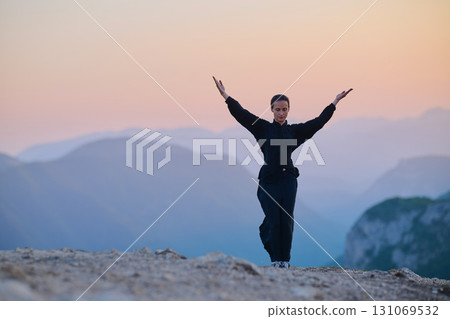 Woman practicing Tai Chi in traditional black outfit on mountain at sunrise, arms outstretched in meditation pose focusing on balance, energy, and mindfulness in nature. Woman practicing Tai Chi in traditional black outfit on mountain at sunrise, arms outstretched in meditation pose focusing on balance, energy, and mindfulness in nature. 131069532