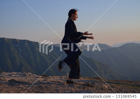 Woman practicing Tai Chi in traditional black outfit on mountain at sunrise, arms outstretched in meditation pose focusing on balance, energy, and mindfulness in nature. 131069545