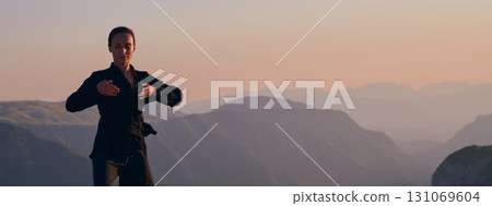 Woman practicing Tai Chi in traditional black outfit on mountain at sunrise, arms outstretched in meditation pose focusing on balance, energy, and mindfulness in nature. 131069604
