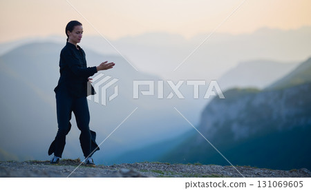 Woman practicing Tai Chi in traditional black outfit on mountain at sunrise, arms outstretched in meditation pose focusing on balance, energy, and mindfulness in nature. 131069605